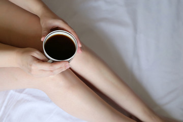Slim girl sitting with coffee cup on the bed, top view. Coffee diet, sexy woman with naked legs and perfect smooth skin, concept of enjoying morning, relaxing at home, weekend