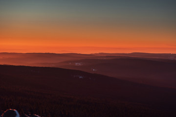 Colorful view from the mountains down to the valley with mountain silhouettes in the distance with moody sunset orange light in winter snow landscape. Torfhaus. Harz Mountains National Park in Germany