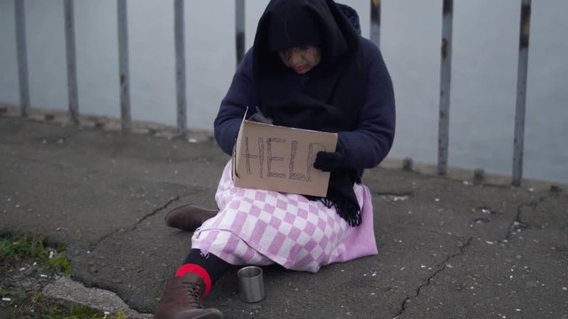 Adult homeless woman sits on the bridge in cold windy grey weather asking for alms and help and writs something on carton