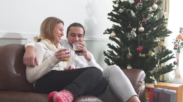 Young guy hugging his girlfriend sitting on a couch drinking coffee next to the Christmas tree. Love relationship.