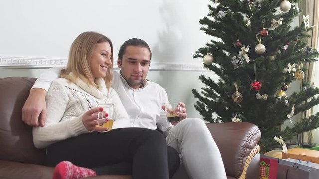 Handsome young guy hugging his beautiful girlfriend sitting on a couch drinking coffee next to the Christmas tree. Love relationship.