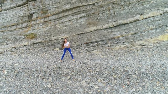 Woman is doing squats the bodyflex during breathing exercises on the sea and rock background. Aerial beautiful footage view.