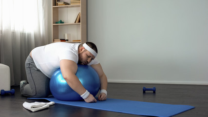 Chubby man resting after workout, lying on fitness ball, desire to lose weight