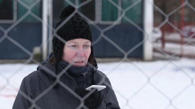 Disappointed woman talking on mobile phone on street behind the chain-link fence that symbolizes obstacle on cold winter day, selective focus