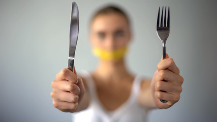 Girl holding fork and knife, mouth closed with tape, food restriction, dieting