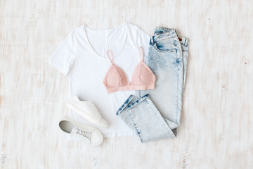 White t-shirt, light blue ripped jeans, pink lace triangle bra, white sneakers on white wooden background. Overhead view of woman's casual outfit. Simple basic women clothes. Flat lay, top view.