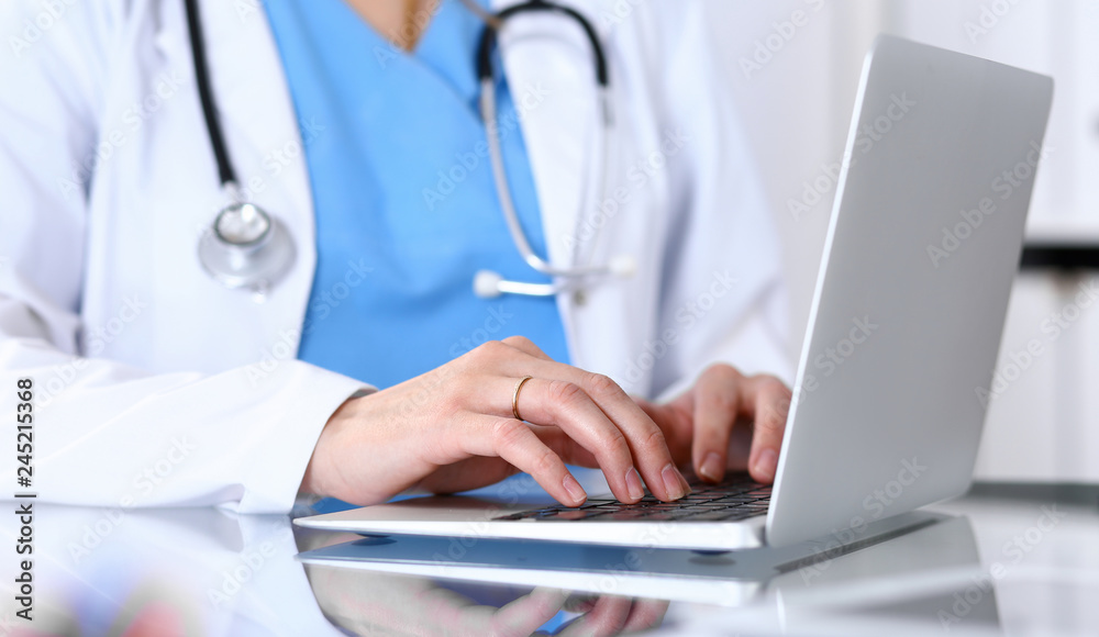 Wall mural Doctor typing on laptop computer while sitting at the glass desk in hospital office. Physician at work. Medicine and healthcare concept