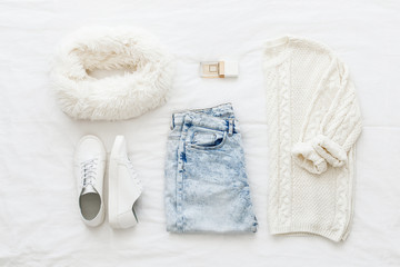 Blue jeans, white knitted sweater, sneakers, fur scarf, perfume lying on bed on white sheet. Overhead view of woman's casual day outfit. Trendy hipster look.  Women winter clothes. Flat lay, top view.