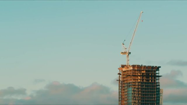 Timelapse of a single under construction building with a crane on top at sunset. The solo building is unfinished and has an elevator move up the side. The sun sets going to darkness. 
