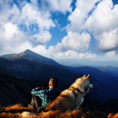 Alone tourist sitting with Alaskan Malamute dog in mountains. Man with his dog on mountain top. Hiker with dog looking at beautiful view in mountains