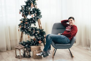 Fototapeta premium A cheerful guy in a red sweater sits near a decorative tree