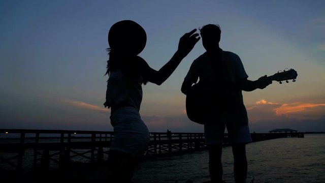 Silhouette of young happy couple having fun with guitar on their beach holiday at sunset