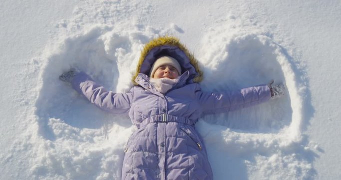 Smiling Kid Laying Down on the Snow Making Snow Angel Shot from Top View on Sunny Day High Angle in Slow Motion Moving Arms and Legs Lying on the Back Close Up