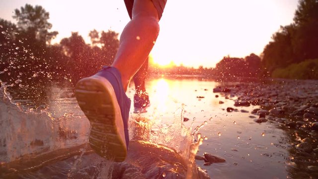 SUPER SLOW MOTION, CLOSE UP, SUN FLARE: Golden evening sunbeams shine on athletic young man jogging along the tranquil river. Picturesque serene nature surrounds sportsman running in the shallow water