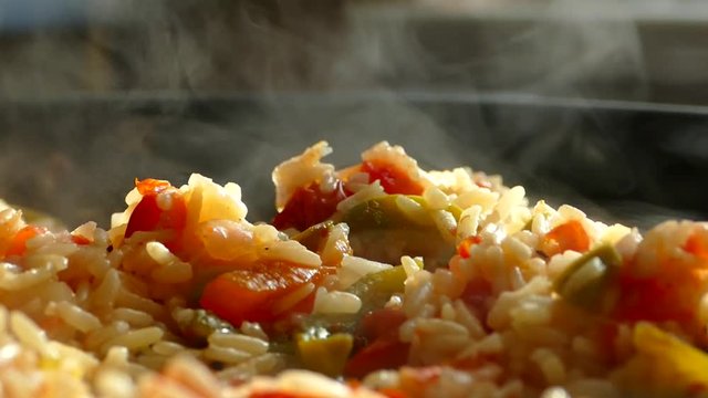 Prepare pilaf in a large frying pan. A dish of rice and pieces of vegetables. Steam comes from a hot lunch. Home kitchen. Pilaf is stirred with a wooden spatula.