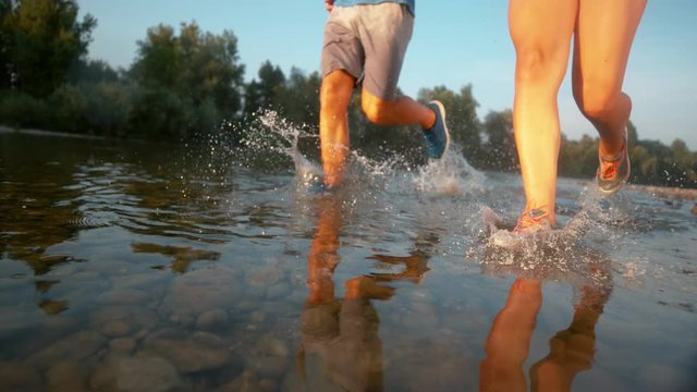 SUPER SLOW MOTION, LOW ANGLE: Athletic couple running in shallow river water on sunny day. Fit young training partners going for a relaxing jog through the forest and along the refreshing stream.