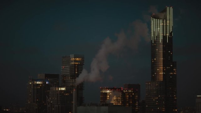 City skyscrapers at night with lights on and smoke stack blowing smoke. Dark and moody with an epic feeling. Downtown big office buildings in a major city. 
