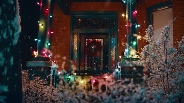 Snow falling outside of house with Christmas lights and a wreath on the front door. 