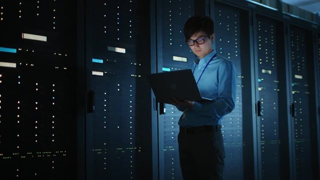 In Dark Data Center: Male IT Specialist Walks along the Row of Operational Server Racks, Uses Laptop for Maintenance. Concept for Cloud Computing, Artificial Intelligence, Supercomputer, Cybersecurity