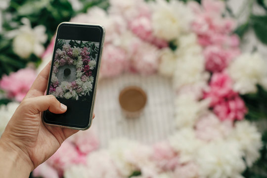 Hand holding phone and taking photo of peonies and coffee cup flat lay on rustic table cloth. Space for text. Blogging and instagram concept. Coffee in glass in peony frame top view on table