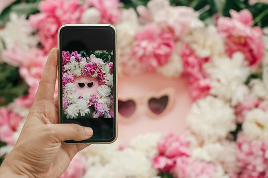 Instagram blogging concept. Hand holding phone and taking photo of stylish girly flat lay of heart shape pink sunglasses with pink and white peonies on pastel pink paper. Hello summer