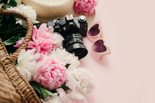 Summer vacation concept. Stylish  pink peonies in straw bag, photo camera, retro sunglasses, hat on pastel pink paper with copy space. International Women's Day. Hello spring