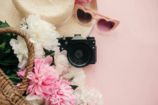 Stylish  flat lay with pink peonies in straw bag, hat, photo camera, retro sunglasses on pastel pink paper with copy space. International Women's Day. Hello spring. Summer vacation