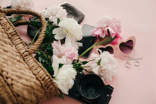 Hello spring image. Stylish  pink peonies in straw bag, phone, photo camera, purse, retro sunglasses on pastel pink paper with copy space. International Women's Day. Summer vacation