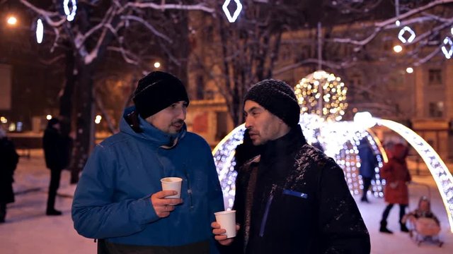 Two men drink coffee in the winter evening in the park in the winter near beautiful Christmas decorations