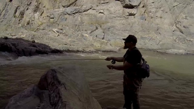 A fly fisherman is silhouetted as he does a simple cast standing in a roaring river.