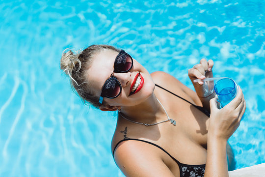 Young beautiful blonde woman in sunglasses with red lips make-up posing in a pool of blue water with a cocktail. Outdoor portrait close up. 