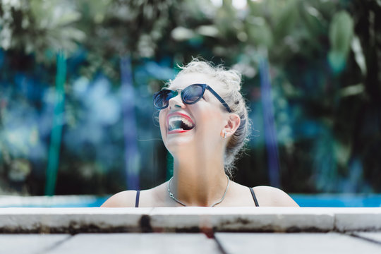 Blurred background of aromatic sticks with young beautiful laughing blonde woman in sunglasses with a good figure with red lips make-up posing in a pool of blue water. Outdoor portrait close up. 