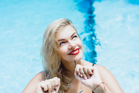 Young beautiful blonde woman with sunglasses with a good figure with red lips make-up posing in a pool of blue water. Outdoor portrait close up. 