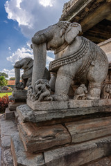 Belavadi, Karnataka, India - November 2, 2013: Veera Narayana Temple. Closeup of two gray stone large statues of elephant on steps to sanctuary. Red roofs of neighboring houses under cloudscape. green