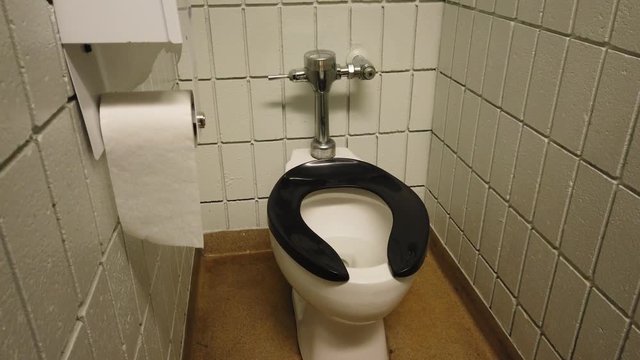 Public washroom toilet stall with white brick walls, silver toilet paper holder and plumbing and a black toilet seat.