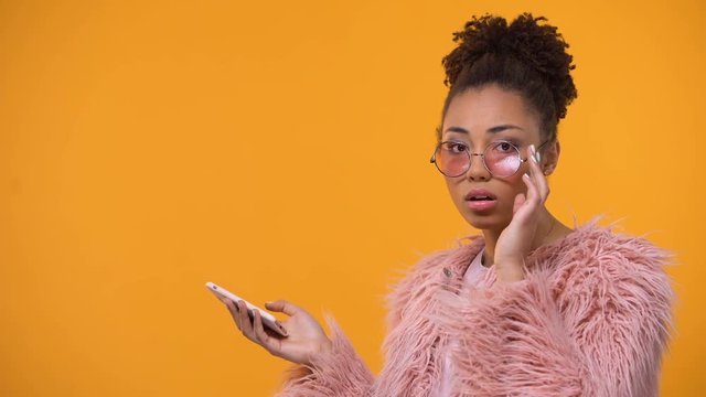 Glamorous girl scrolling web-pages on smartphone, isolated yellow background