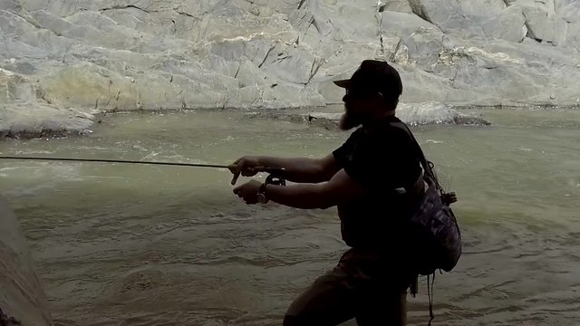 A fly fisherman is silhouetted against a roaring river while mending line in slow motion.