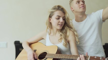 Young couple is sitting on the bed in their room. Woman is playing the guitar and singing and man is hugging her and making a selfie of two of them.