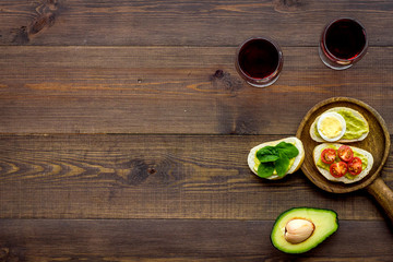 Obraz na płótnie Canvas Light appetizer with avocado. Toasts with vegetables and guacamole near glasses of wine on dark wooden background top view space for text