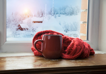 Winter background - cup with candy cane, woolen scarf and gloves on windowsill and winter scene outdoors. Still life with concept of spending winter time at cozy home with cold weather outdoors
