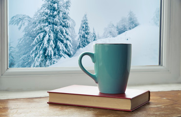 Winter background - cup with candy cane, woolen scarf and gloves on windowsill and winter scene outdoors. Still life with concept of spending winter time at cozy home with cold weather outdoors