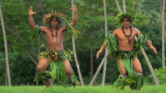 Polynesian young men in grass skirts with flower headdress dancing hula war dance while entertaining barefoot outdoors Tahiti French Polynesia 