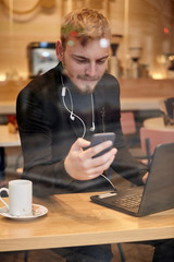 one young man, sitting in coffee shop and using his smart-phone, with laptop and cup nearby. Shoot thought window outside (with reflections).