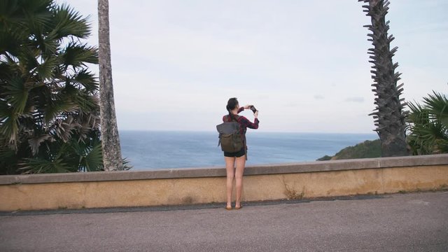 Woman traveler with backpack admiring sea view and making photo on the shore of a tropical beach sunset using smartphone. Female wearing red checkered shirt and sunglasses.