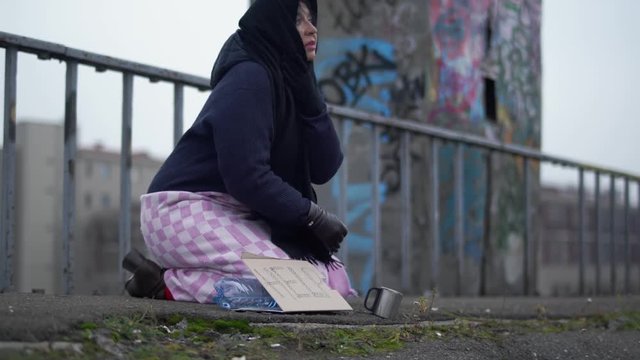 Elderly homeless woman with outstretched hand sits on the bridge in cold windy grey weather asking for alms and help and one woman stopped to put coins