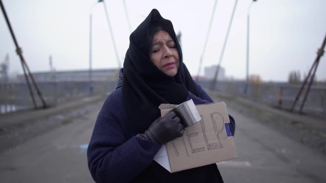 Portrait of adult sad homeless woman stay on the bridge in cold windy grey weather asking for alms and help