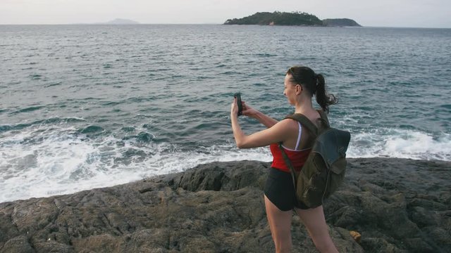 Woman traveler with backpack admiring sea view and making photo on the shore of a tropical beach sunset using smartphone. Female wearing red checkered shirt and sunglasses.