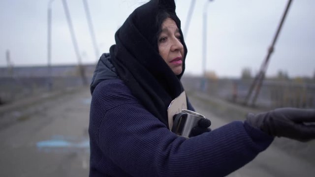 Woman gives alms to a beggar standing on the bridge in cold windy grey weather asking for alms and and charity