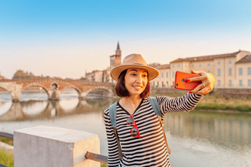 Smiling traveler woman taking selfie on her smartphone at the background of the Verona Bridge Ponte...