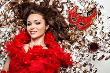 Charming young woman lying on the floor among golden tinsel, smiling close-up next to a red mask and glass of wine